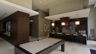 This single-floor space is a masterfully designed entertainment venue that blends modern aesthetics with functional luxury. The open-concept layout centers around a full-size billiards table, finished in rich dark wood tones that complement the room's contemporary design palette. Elevated ceilings with recessed lighting create a sense of openness and volume, while the neutral warm tones of the walls and light tile flooring provide a refined, cohesive backdrop throughout.

Adjacent to the billiards area is a well-appointed wet bar and kitchenette, anchored by a stone tile accent wall that adds texture and visual depth. Dark cabinetry pairs elegantly with a light countertop surface, and a row of bar seating invites casual dining and socializing. Pendant lights hang above the bar area, casting warm, focused illumination that enhances the lounge-like ambiance.

A wall-mounted flat-screen television is integrated seamlessly into the bar area, making it an ideal setting for entertaining. Framed artwork adorns the walls, adding a curated, polished touch. A stainless steel refrigerator provides practical functionality within the stylish kitchenette. The overall design of the space is sophisticated and intentional, offering a premium environment well-suited for social gatherings and recreational use.