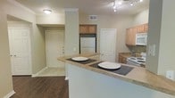 Located at 1420 McDermott Dr. in Allen, Texas, Settler's Gate Apartment presents a thoughtfully designed single-floor residence that balances comfort and functionality. The open-concept kitchen serves as the heart of the home, showcasing light wood cabinetry, laminate countertops, and a full suite of appliances including a refrigerator and electric range. A peninsula breakfast bar with seating for two creates a natural gathering spot, making the kitchen ideal for both casual dining and entertaining. Track lighting overhead provides focused illumination throughout the cooking area, while crown molding adds a refined architectural touch to the space. The flooring transitions seamlessly from rich, dark hardwood in the main living areas to classic tile in the entryway, creating clear visual zones throughout. The entryway is bright and welcoming, featuring a solid panel front door with multiple security locks for added peace of mind. Neutral wall tones throughout the residence create a clean, versatile backdrop that complements a variety of décor styles. The overall ambiance is warm and inviting, with a practical layout that maximizes the use of available space while maintaining an open, airy feel.