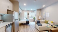 This efficient studio unit is designed to maximize functionality within a single open-plan floor. The main living area seamlessly integrates sleeping, lounging, and living zones, anchored by light wood-look flooring and a neutral color palette that lends a bright, airy ambiance throughout. A Murphy bed folds neatly against the wall, flanked by built-in overhead cabinetry that provides ample storage without sacrificing floor space. Large windows fitted with roller shades allow natural light to fill the room while offering privacy, and views of the surrounding urban streetscape are visible from the sleeping area. A comfortable sofa and side table define the living zone, while decorative wall art and an easel add a creative, personalized touch.

The kitchen is compact yet fully equipped, featuring stainless steel appliances including an electric range, over-the-range microwave, dishwasher, and refrigerator. Light wood cabinetry extends from counter height to the ceiling, maximizing storage, while a white penny-tile backsplash adds a subtle design detail. The bathroom is clean and minimal, finished in all-white tile with a walk-in shower enclosed by a curtain. Recessed lighting throughout the unit maintains a polished, modern feel.