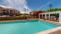 Located at 3600 Corriere Rd in Easton, Pennsylvania, this outdoor amenity space is designed to deliver a resort-style experience for its residents. At the heart of the space is a generously sized swimming pool with sparkling blue water, accented by a decorative tiled logo on the pool floor and a shallow wading entry area. The pool is surrounded by an expansive, warm-toned paved sundeck furnished with lounge chairs and shaded dining areas outfitted with green market umbrellas, providing ample space for both relaxation and socializing.

A prominent white pergola lines one side of the pool deck, offering a sheltered corridor that adds architectural character and partial shade. Lush landscaping, including flowering lavender and ornamental trees, frames the area and enhances its inviting atmosphere. Stone retaining walls and decorative fencing define the perimeter, creating a sense of enclosure while maintaining open views of the surrounding residential buildings.

Adjacent to the pool, a single-story clubhouse with a pitched shingle roof and expansive windows provides a bright indoor retreat that seamlessly connects to the outdoor deck. The overall ambiance is vibrant and welcoming, ideal for outdoor gatherings under open skies in a well-appointed community setting.