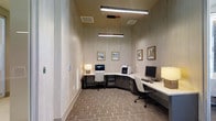 This single-floor business center is thoughtfully designed to provide a functional and comfortable workspace for guests or occupants seeking productivity. The room features an L-shaped, dark-toned workstation that wraps along two walls, maximizing the available workspace while maintaining an open feel. Multiple desktop computers and a multifunction printer are conveniently arranged along the countertop, making the space fully equipped for a variety of office tasks. Each workstation is paired with a modern white ergonomic chair, offering comfort during extended use. Soft, warm lighting is provided by sleek fluorescent overhead fixtures and accent desk lamps, creating a well-lit yet inviting atmosphere. The walls are finished with a subtle textured wallcovering in neutral tones, lending the space a clean and professional aesthetic. A collection of framed artwork adorns the walls, adding a polished and welcoming touch to what might otherwise be a purely utilitarian room. The flooring features a stone-patterned tile laid in an earthy palette, contributing to the room's grounded, sophisticated character. Ceiling-mounted HVAC vents ensure a comfortable climate throughout. Overall, this business center balances practicality with refined design, making it a well-appointed space for work-related needs.