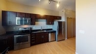 Located at 2900 DeMers Ave in Grand Forks, North Dakota, this single-floor unit offers a thoughtfully designed layout with modern finishes throughout. The entry hallway features light hardwood-style flooring and a sliding wood-panel door, leading seamlessly into the main living areas.

The kitchen is a standout feature, outfitted with rich dark-wood cabinetry, dark granite-style countertops, and a full suite of stainless steel appliances including a refrigerator, electric range, built-in microwave, and dishwasher. Track lighting overhead enhances the clean, contemporary feel. A window at the end of the kitchen brings in natural light, making the space feel bright and functional.

The living area is open and airy, with the same hardwood-style flooring continuing throughout. Neutral-toned walls and a window allow for natural light, creating a comfortable and inviting atmosphere.

The bedroom offers a carpeted floor and a quiet, understated feel with warm neutral wall tones and a ceiling light fixture. An in-unit washer is also conveniently located within the unit's layout.

The bathroom includes a full soaking tub and shower combination, a dark-cabinetry vanity with granite-style countertop, and built-in shelving for additional storage. Light tile flooring and a drop ceiling complete the clean, practical design.