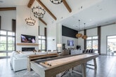 The Laurel at Dry Creek clubhouse in Huntsville, Alabama presents a thoughtfully designed single-story community space that blends modern farmhouse aesthetics with upscale finishes throughout. A striking geometric parquet-style wood-patterned floor runs continuously across the facility, serving as a unifying design element that anchors each distinct zone within the open layout.

The reception and leasing area features a large communal table surrounded by plush upholstered chairs, suspended beneath a trio of cone-shaped pendant lights. Wood-clad structural columns add warmth and texture, while panel-molded walls in soft neutral tones display curated artwork, contributing to a refined yet approachable atmosphere.

Adjacent spaces include a well-appointed lounge area with soaring ceilings supported by exposed wooden beams and dramatic floor-to-ceiling windows that flood the interior with natural light and offer expansive outdoor views. A white-brick fireplace, chandelier lighting, and a shuffleboard table enhance the relaxed, social character of this area.

A dedicated game and entertainment room features additional shuffleboard and billiards tables, a media wall with a mounted television, and a textured accent wall. Throughout the clubhouse, a carefully curated mix of pendant and chandelier fixtures, neutral furnishings, and natural materials creates a cohesive, inviting ambiance suited for both casual gatherings and community events.
