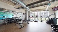 Located at 1601 Central Ave in Saint Petersburg, Florida, this single-floor fitness center offers a well-appointed and thoughtfully designed workout facility. The space embraces an industrial-modern aesthetic, highlighted by exposed concrete ceilings with prominent silver ductwork running overhead, adding a distinctive urban character to the environment.

The main workout area is generously proportioned, featuring warm wood-tone flooring throughout that contrasts effectively with the raw ceiling elements. A wall of large, floor-to-ceiling windows floods the cardio zone with abundant natural light while offering views of the surrounding streetscape, creating an energizing atmosphere. This area is equipped with multiple treadmills, upright bikes, and a stair climber, all positioned to take advantage of the outdoor views.

The free weights section includes a full dumbbell rack accompanied by adjustable benches and wall-mounted mirrors, ideal for strength training. Additional areas house cable and resistance machines, stability balls, and a mini trampoline for functional fitness. Glass-walled rooms with black steel-framed doors provide semi-private spaces that open from the main floor, one appearing to serve as a lounge or meeting area, and another as a dedicated strength training zone. A small refreshment and sanitation station is also conveniently located within the facility.