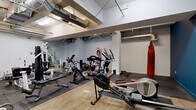 This fitness facility offers a comprehensive workout environment housed within a single open floor. The space features an industrial-inspired aesthetic, with exposed ductwork and ventilation systems running along the ceiling, complemented by a combination of polished concrete and rubber mat flooring throughout the exercise areas.

The main workout area is divided into distinct functional zones. The cardio section is well-stocked with a variety of equipment, including treadmills, stationary bikes, elliptical trainers, and a rowing machine, providing ample options for cardiovascular training. A dedicated strength training zone features a multi-function cable machine, weight benches, barbells, and a rack of dumbbells, catering to a full range of resistance exercises.

A striking red punching bag hangs prominently in one section of the gym, establishing a dedicated boxing and combat training area. Large mirrors are mounted along select walls, aiding in form correction and visually expanding the space. A wall-mounted television is also present in the strength training area for entertainment during workouts.

The walls are painted in a two-tone scheme, with clean white in the main area transitioning to a bold teal-blue accent wall in a secondary section, adding visual energy to the environment. A solid wood entry door with a transom window provides access to the space, adding a warm, classic contrast to the otherwise utilitarian design.