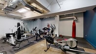 This fitness facility offers a comprehensive workout environment housed within a single open floor. The space features an industrial-inspired aesthetic, with exposed ductwork and ventilation systems running along the ceiling, complemented by a combination of polished concrete and rubber mat flooring throughout the exercise areas.

The main workout area is divided into distinct functional zones. The cardio section is well-stocked with a variety of equipment, including treadmills, stationary bikes, elliptical trainers, and a rowing machine, providing ample options for cardiovascular training. A dedicated strength training zone features a multi-function cable machine, weight benches, barbells, and a rack of dumbbells, catering to a full range of resistance exercises.

A striking red punching bag hangs prominently in one section of the gym, establishing a dedicated boxing and combat training area. Large mirrors are mounted along select walls, aiding in form correction and visually expanding the space. A wall-mounted television is also present in the strength training area for entertainment during workouts.

The walls are painted in a two-tone scheme, with clean white in the main area transitioning to a bold teal-blue accent wall in a secondary section, adding visual energy to the environment. A solid wood entry door with a transom window provides access to the space, adding a warm, classic contrast to the otherwise utilitarian design.