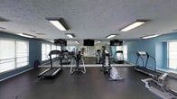 This fitness center offers a practical and well-organized space for cardiovascular and general exercise. The room is furnished with a selection of cardio equipment, including treadmills, elliptical trainers, and a stationary bike, arranged to allow comfortable movement between machines. A full-length mirrored wall spans one side of the room, visually expanding the space while allowing users to monitor their form during workouts. Multiple flat-screen televisions are mounted at eye level, providing entertainment options during exercise sessions. The flooring consists of dark rubber matting, a durable and practical surface well-suited for a fitness environment. Natural light enters through windows fitted with horizontal blinds on opposing walls, contributing to a bright and energizing atmosphere. Overhead fluorescent lighting provides consistent illumination throughout the space. The walls are painted in a cool blue-gray tone, lending the room a clean and focused aesthetic. The textured ceiling adds a subtle architectural detail. Overall, the fitness center is a functional, well-lit space designed to meet everyday exercise needs.