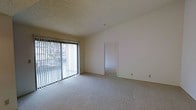 This single-floor space presents a clean, functional layout with neutral finishes throughout. The main living area is carpeted in a light, neutral tone that extends seamlessly into the adjoining room visible through an open doorway, creating a cohesive flow between spaces. A textured popcorn ceiling adds dimension overhead, while smooth, white-painted walls provide a bright, open backdrop throughout.

A prominent sliding glass door fitted with vertical blinds dominates one wall of the main living area, allowing natural light to filter into the space and offering a view of an outdoor area with visible greenery. The blinds provide adjustable light control, contributing to the room's versatility. The sliding door also serves as direct access to the exterior.

The open doorway on the opposite wall leads to a secondary carpeted room, maintaining the same neutral palette and consistent design language. The overall ambiance is understated and practical, characterized by simple architectural details and a move-in-ready, blank-canvas quality that allows for flexible furnishing and personalization.