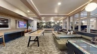 Located at 1604 W Lincoln Ave in Anaheim, California, this generously sized single-floor community lounge and entertainment space is thoughtfully designed for socializing and recreation. The open layout is anchored by a striking botanical mural wall that adds a bold, artistic focal point to the room, complemented by sophisticated patterned carpeting that flows throughout the main gathering areas.

The space is divided into distinct yet seamlessly connected zones. A plush lounge area features tufted leather sectional seating arranged around low wood coffee tables, ideal for relaxed conversation. Adjacent to it, a dedicated game area includes shuffleboard and foosball tables, encouraging friendly competition. A built-in bar counter with a textured tile backsplash and bar-height seating faces multiple wall-mounted flat-screen TVs, creating a sports lounge atmosphere. A separate alcove houses a racing simulator station set against large-format tile flooring, providing an immersive gaming experience.

Overhead, a coffered ceiling with warm cove lighting and globe pendant fixtures creates a layered, inviting ambiance. A mirrored partition wall adds depth and light to the space. Large windows along one side invite natural light, while recessed lighting ensures consistent illumination throughout. The overall design blends modern comfort with entertainment-forward functionality.