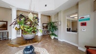 This well-appointed single-story community clubhouse offers a thoughtfully designed layout that balances elegance with functionality. The welcoming lobby greets visitors with decorative white columns, warm neutral tones, and lush floral arrangements atop a herringbone-patterned tile floor, creating a refined first impression. Adjacent to the entry, a staffed reception area provides convenient access to building services.

The central lounge area is a standout feature, highlighted by built-in white display shelving, crown molding, and comfortable seating arranged around a classic round coffee table atop a vibrant area rug. A stone accent fireplace wall with a mounted flat-screen television anchors a cozy sitting nook nearby.

The dining area is bathed in natural light from floor-to-ceiling windows and glass doors, dressed with elegant drapes and wood blinds that frame outdoor views. A rich dark wood dining table and chairs complete the space.

The expansive business center offers multiple computer workstations with granite countertops, hardwood-style flooring, a tray ceiling, and ample printing equipment, providing a fully functional workspace for residents.

The kitchen is equipped with dark granite countertops, rich wood cabinetry, stainless steel appliances including a refrigerator and range, and a large center island with pendant lighting, making it ideal for community gatherings and events.