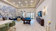 This thoughtfully designed single-floor amenity space blends sophisticated style with social functionality. The main area is anchored by a full-size pool table with a bold blue felt surface, set atop light wood-look flooring that runs throughout. A tray ceiling with warm LED accent lighting adds architectural depth, while recessed lighting ensures the space is well-illuminated throughout.

One wall showcases a striking large-scale floral mural rendered on exposed white brick, serving as a dramatic artistic focal point. Opposite, a white brick accent wall hosts a wall-mounted flat-screen TV flanked by decorative gold sconce lighting, above a richly detailed wood media console. A generously sized navy sectional sofa, adorned with an assortment of green and patterned throw pillows, creates a comfortable lounge area beneath framed artwork.

Floor-to-ceiling French doors flood the space with natural light and provide outdoor access, dressed with navy and white drapes. A dedicated wet bar area features an L-shaped quartz countertop, warm wood cabinetry, a beverage refrigerator, and a wall-mounted TV—ideal for hosting. The overall ambiance is vibrant yet refined, combining coastal-inspired tones of navy, teal, and gold for a welcoming, upscale entertaining environment.