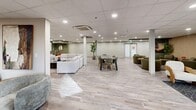 This generously sized social lounge in Calgary, Alberta, offers a thoughtfully designed communal space spread across a single open floor. The layout is organized into several distinct seating zones, each with its own character and atmosphere, yet unified by light wood-tone flooring, neutral walls, and consistent recessed lighting throughout the drop-panel ceiling.

One area is anchored by a striking floor-to-ceiling mountain landscape mural in warm earthy tones, complemented by rich brown leather sofas, a solid wood coffee table, and a vertical slat accent wall — creating a cozy, nature-inspired retreat. Adjacent to this, a bright nook features a serene forest mural as its backdrop, paired with a white sofa, curved armchairs, and a patterned area rug beside a window with outdoor views.

A lounge zone with rounded, upholstered barrel chairs in soft grey surrounds a linear electric fireplace set into the wall, with potted trees and framed artwork adding organic warmth to the space. The central open area accommodates a large sectional sofa arrangement and a dedicated dining table with chairs, providing functional space for both casual socializing and group gatherings.

Throughout, lush indoor plants, layered rugs, and carefully curated furnishings contribute to an inviting, modern ambiance suited for community use.