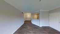 This single-floor residence showcases an open-concept design that connects the living area, dining space, and kitchen in a cohesive, flowing layout. Dark hardwood-style flooring extends throughout, providing a rich contrast against the bright white walls and crisp crown molding that frames each room. The living area is spacious and well-proportioned, with neutral-toned walls and clean architectural lines that create a welcoming, move-in-ready atmosphere.

A half-wall partition defines the kitchen pass-through, which features light countertops and visibility into the main living space, making it ideal for entertaining. The kitchen is equipped with stainless steel appliances and benefits from recessed lighting overhead. Beyond the kitchen, the dining area is distinguished by elegant wainscoting panels along the lower walls and an overhead chandelier that adds a refined, classic touch to the space.

Throughout the unit, white baseboards and trim work provide polished finishing details, while neutral paint tones contribute to a bright, airy ambiance. The consistent flooring and cohesive color palette tie each zone together seamlessly, resulting in a well-designed, functional living environment.
