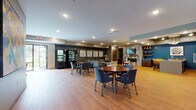 This well-appointed single-floor amenity space offers a thoughtfully designed environment for gathering, relaxing, and socializing. The entrance lobby sets an inviting tone with large-format tile flooring, floor-to-ceiling glass doors, and eye-catching sculptural pendant light fixtures that create a warm first impression. A drop box and adjacent offices are conveniently accessible from the entry corridor.

The open common area is anchored by a generously sized lounge featuring a large sectional sofa, a patterned blue area rug, and a stylish coffee table, all set against a dramatic textured chevron accent wall. Framed artwork adds color and character throughout. A stone fireplace with built-in shelving and a wall-mounted TV creates a cozy focal point on one end, while vending machines offer added convenience nearby.

Multiple dining areas accommodate both casual and group seating, with round and rectangular tables paired with blue and red chairs atop warm wood-look flooring. The kitchenette is sleekly finished with rich wood cabinetry, quartz countertops, subway tile backsplash, and stainless steel appliances, highlighted by a bold teal accent wall.

A secondary seating nook with red upholstered chairs and colorful artwork provides an additional lounge area, rounding out this versatile and vibrant community space.