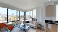 This single-floor residence showcases a thoughtfully designed living area that prioritizes natural light and urban connectivity. The standout feature is the dramatic corner window configuration, where expansive floor-to-ceiling black-framed windows meet at an angle, flooding the interior with abundant daylight while framing panoramic views of the surrounding cityscape, including tree-lined streets and the broader urban skyline beyond.

The living room is finished with clean white walls and smooth ceilings that amplify the sense of openness. Light hardwood flooring runs throughout, adding warmth to the otherwise crisp, contemporary palette. Horizontal window blinds provide adjustable light control without compromising the sweeping views.

The space is furnished with a neutral-toned sofa accented by navy and gold throw pillows, complemented by a glass-topped coffee table with brass-finished legs — details that reinforce the modern aesthetic. A minimalist side table and subtle pendant lighting complete the refined, curated atmosphere.

Overall, the residence exudes a bright, sophisticated ambiance, well-suited for those who appreciate contemporary design and elevated urban living.