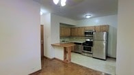 This single-floor space showcases an open-concept kitchen that serves as the focal point of the layout. The kitchen is outfitted with a full suite of stainless steel appliances, including a gas range with oven, an over-the-range microwave, and a full-size refrigerator, all complemented by warm-toned wood cabinetry that extends to the upper walls for generous storage. A decorative tile backsplash runs along the countertop, adding a subtle design detail to the cooking area. A peninsula-style breakfast bar with a raised wooden counter extends into the adjacent living area, creating a natural divide between spaces while offering additional seating and prep surface. The kitchen floor features light-toned tile, while the adjoining living area transitions to classic parquet hardwood flooring, lending warmth and character to the space. Recessed and flush-mount ceiling light fixtures provide practical overhead illumination throughout. A door visible along one wall suggests convenient access to additional rooms or building entry. The overall layout is functional and straightforward, designed to maximize usability within a compact, well-organized single-floor footprint.