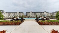 This outdoor amenity space is designed to deliver a resort-like experience, centered around a generously sized swimming pool flanked by an expansive paved sundeck. The pool area is lined with rows of dark-framed chaise lounges and accent lighting pillars, creating a refined and inviting atmosphere for relaxation. Lush landscaping with ornamental grasses and shrubs borders the deck, adding color and texture to the hardscape surroundings.

Adjacent to the pool, a covered outdoor lounge patio serves as a standout architectural feature. Soaring vaulted ceilings are clad in warm natural wood, supported by exposed timber beams and complemented by ceiling fans that ensure comfort year-round. The structure is built with striking white limestone masonry columns that lend a sense of permanence and craftsmanship. Floor-to-ceiling windows and large glass doors line the interior-facing wall, allowing views into the clubhouse while flooding the covered area with natural light.

The lounge patio is furnished with dark wicker sectional seating and accent chairs upholstered in bold cobalt blue, creating a stylish and comfortable outdoor living area. The combination of natural stone, warm wood tones, and modern furnishings gives this space a balanced aesthetic that is both sophisticated and welcoming. The surrounding multi-story residential buildings are visible in the background, providing a sense of the broader community context.