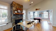 The Edge at Kutztown is a dynamic, single-floor community and coworking space designed to foster collaboration, recreation, and productivity. The main area greets visitors with soaring vaulted ceilings, warm hardwood flooring, and an abundance of natural light streaming through multiple windows and a overhead skylight. A dramatic floor-to-ceiling stone fireplace serves as the centerpiece of the lounge area, complete with a wooden mantel and seasonal décor, lending the space a welcoming, residential warmth. Flexible workstations with rolling tables and mixed seating accommodate individual and group work throughout the open layout. A dedicated game room, partially enclosed with a pass-through opening, features a full-size pool table, ping pong table, and university pennants that nod to the nearby Kutztown University community. A built-in bar-height counter with stools provides a casual refreshment and hangout station, while a large chalkboard wall adds an interactive, creative element to the space. The well-lit entry vestibule offers a welcoming first impression with glass double doors and branded signage. A private office or meeting room with carpet and darker furnishings provides a quieter retreat. Together, the varied zones make this a well-rounded, energetic hub suited for work, socializing, and leisure.