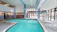 This indoor pool facility offers a well-appointed aquatic environment designed for year-round enjoyment. The centerpiece is a generously sized rectangular swimming pool with crystal-clear turquoise water, framed by clean tile surrounds and clearly marked lane depths. The space is characterized by soaring ceilings finished in white paneling, supported by light blue columns that lend a cohesive and refreshing color palette throughout. Large floor-to-ceiling windows and a glass-paneled wall along one side of the facility flood the area with natural light, providing views of the surrounding exterior and creating an open, airy atmosphere. Lounge chairs and seating areas are positioned along the pool deck, offering comfortable spots to relax before or after a swim. A hot tub or spa feature is also visible along one edge of the pool, providing an additional relaxation option. The overall ambiance is clean, bright, and welcoming, with the blue-accented architecture and reflective water surface contributing to a calm and resort-like setting. The facility appears well-maintained, making it a functional and visually appealing space for leisure and recreation.