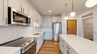This thoughtfully designed two-bedroom, two-bathroom residence offers a cohesive blend of modern aesthetics and practical functionality across a single floor. The kitchen is a standout feature, showcasing white shaker cabinetry with matte black hardware, quartz countertops, a decorative lattice tile backsplash, and a full suite of stainless steel appliances including a range, microwave, dishwasher, and refrigerator. A central island provides additional prep space and casual seating, while globe pendant lights overhead add a contemporary touch. The kitchen flows seamlessly into the living area, which is brightened by a large sliding glass door that opens onto a private balcony with outdoor views of surrounding greenery. Hardwood-style flooring runs throughout the main living areas, lending warmth and continuity. The primary bedroom offers plush carpeting, a generous walk-in closet with built-in shelving, and a dedicated en-suite bathroom featuring a white vanity, a large framed mirror, and a glass-enclosed shower. The second bedroom is equally comfortable, with carpeted flooring and a sizable window that fills the room with natural light. A second full bathroom serves the remaining spaces. A dedicated laundry area houses a stacked full-size washer and dryer unit alongside the water heater and HVAC system, offering added convenience within the home.