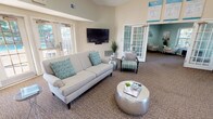 This Clubhouse and Business Center is a well-appointed, single-floor community facility designed to serve both social and professional needs. The main lounge area makes an immediate impression with soaring vaulted ceilings, dual ceiling fans, and a dramatic arched transom window above the entrance that floods the space with natural light. Neutral warm tones on the walls are complemented by accents of teal and seafoam green throughout the furnishings and décor, creating a fresh, coastal-inspired ambiance. Comfortable seating arrangements include a plush sofa, accent chairs, and a stylish metallic coffee table, all oriented around a wall-mounted flat-screen television. French door access leads directly to an outdoor pool area, framing inviting views of the surrounding landscape. A dedicated business center nook, tucked within its own room accessed through a classic glass-paned door, offers a computer workstation with printer and ample natural light through slatted blinds. A separate lounge room with soft blue-green walls provides a quieter seating retreat with decorative wall art. The facility also includes a kitchenette with dark cabinetry, a stainless steel refrigerator, tiled backsplash, and a breakfast bar with seating. A welcoming reception area with a front desk and entrance rug completes this versatile community space.