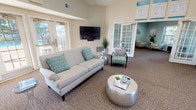 This Clubhouse and Business Center is a well-appointed, single-floor community facility designed to serve both social and professional needs. The main lounge area makes an immediate impression with soaring vaulted ceilings, dual ceiling fans, and a dramatic arched transom window above the entrance that floods the space with natural light. Neutral warm tones on the walls are complemented by accents of teal and seafoam green throughout the furnishings and décor, creating a fresh, coastal-inspired ambiance. Comfortable seating arrangements include a plush sofa, accent chairs, and a stylish metallic coffee table, all oriented around a wall-mounted flat-screen television. French door access leads directly to an outdoor pool area, framing inviting views of the surrounding landscape. A dedicated business center nook, tucked within its own room accessed through a classic glass-paned door, offers a computer workstation with printer and ample natural light through slatted blinds. A separate lounge room with soft blue-green walls provides a quieter seating retreat with decorative wall art. The facility also includes a kitchenette with dark cabinetry, a stainless steel refrigerator, tiled backsplash, and a breakfast bar with seating. A welcoming reception area with a front desk and entrance rug completes this versatile community space.