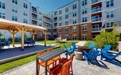 Located at 10945 Nuckols Rd in Glen Allen, Virginia, this two-story residential community presents a thoughtfully designed exterior and communal outdoor environment. The building's architecture combines classic red brick facades with modern grey horizontal siding, creating a visually dynamic aesthetic complemented by private balconies on upper floors.

At the heart of the property lies a spacious central courtyard that serves as the community's primary outdoor gathering area. A wooden pergola with a metal roof anchors one end of the courtyard, providing a shaded dining and seating area. Adjacent to it, colorful Adirondack chairs surround a round fire pit, offering a relaxed social atmosphere. Freestanding hammocks are positioned on the open lawn, inviting leisure and relaxation in the sunlit green space.

Concrete walkways weave throughout the courtyard, connecting various seating areas, planting beds filled with ornamental grasses and shrubs, and ground-floor unit patios enclosed by wrought iron fencing. Lush landscaping lines the building perimeter, adding greenery and privacy to ground-level residences. The courtyard also features a covered lounge area with potted plants and additional seating directly accessible from the building's interior common spaces, blending indoor and outdoor living seamlessly.