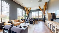Located at 200 Resource Pkwy in Franklin, Tennessee, this single-floor living space delivers a compelling blend of industrial character and refined design. The room is immediately defined by its soaring ceilings, which are anchored by a bold V-shaped arrangement of exposed natural wood beams — a standout architectural feature that draws the eye upward and adds rustic warmth against the deep slate-gray walls.

Expansive floor-to-ceiling arched and gridded windows line the space, inviting abundant natural light and offering open views of the surrounding Franklin skyline and greenery. The warm-toned hardwood flooring runs throughout, grounding the room with continuity and elegance.

The living area is thoughtfully arranged to serve multiple functions. A seating zone features a rich tan leather sofa, navy blue accent chairs with gold detailing, and a round marble-topped coffee table set atop a layered area rug. A dedicated dining area accommodates a wood-top table surrounded by plush velvet chairs with gold-framed legs, accompanied by tall bar-height seating near the windows. On the opposite wall, a built-in entertainment unit with arched cabinetry and glass-front drawers houses a large flat-screen television, flanked by framed artwork. The overall ambiance is sophisticated, airy, and modern with artful industrial touches throughout.