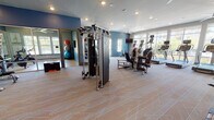 This fitness center in Jacksonville, FL (32246) is a thoughtfully designed, single-floor facility offering a comprehensive workout environment for users of all fitness levels. The space is defined by bold blue accent walls, light-toned patterned flooring, and a recessed lighting system complemented by large ceiling fans that maintain air circulation throughout.

The main workout floor is generously proportioned, providing ample space between equipment for comfortable movement. A dedicated strength training area features a multi-functional cable machine, selectorized weight machines, free weights, kettlebells, medicine balls, and a heavy bag, catering to a full range of resistance and functional training exercises. A mirrored section with open floor space and stability balls supports stretching and balance-focused workouts.

The cardio zone is positioned along a wall of large windows, offering an abundance of natural light and scenic outdoor views of lush landscaping and palm trees. Treadmills, stair climbers, and rowing machines are available, each equipped with individual monitors. Wall-mounted TVs are also present throughout.

Large glass panel doors connect the fitness center to an adjacent common area, enhancing the sense of openness. Motivational artwork adorns the walls, contributing to an energetic yet welcoming ambiance well-suited for a focused and productive workout experience.