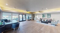 This single-floor community clubhouse is thoughtfully designed to offer a welcoming and functional environment for residents. The space features wide-plank light wood flooring throughout, creating a cohesive and airy aesthetic that ties the various zones together seamlessly.

Upon entry, a professional leasing office greets visitors with a sleek, dark wood L-shaped reception desk positioned in front of large windows fitted with roller shades, allowing ample natural light while maintaining privacy. Dual ceiling fans and recessed lighting are featured prominently, complemented by a distinctive yellow-accented tray ceiling detail that adds a warm, modern touch throughout the space.

The central lounge area is furnished with a contemporary gray sofa, patterned accent chairs, and a patterned area rug, all anchored beneath vibrant teal and green abstract wall art. A kitchenette nearby features white upper cabinetry, slate-blue lower cabinets, a chevron tile backsplash, stainless steel appliances, and under-counter beverage and water bottle refrigerators — ideal for resident gatherings.

Large glass double doors open directly to an outdoor pool area, flooding the interior with natural light and providing convenient indoor-outdoor access. A dedicated fitness area with exercise equipment rounds out the amenities. The overall ambiance is clean, bright, and inviting, reflecting a well-appointed residential community facility.