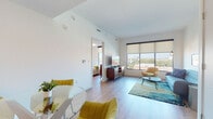 This well-appointed single-floor residence offers a thoughtfully designed layout that maximizes both comfort and functionality. The open-plan living and dining area is bathed in natural light, courtesy of large floor-to-ceiling windows that frame sweeping city views. Light wood-style flooring runs throughout, contributing to a warm, cohesive aesthetic. The living area is generously proportioned, accommodating a seating arrangement centered around a mid-century modern sofa, accent chairs in mustard yellow, and a contemporary coffee table set atop a vibrant area rug.

The kitchen is efficiently laid out with dark lower cabinetry, light upper cabinets, and a textured stone tile backsplash. Stainless steel appliances, including a range, over-the-range microwave, and refrigerator, complete the space alongside sleek dark countertops and a stainless steel sink.

The bedroom continues the light, airy palette with crisp white walls, wood-style flooring, and a window offering pleasant city views. A substantial mid-century dresser provides ample storage alongside the room's generous proportions.

The bathroom features marble-style tile flooring, a full soaking tub with shower and curved rod, light wood cabinetry, and a dark countertop vanity — all finished in a clean, contemporary style throughout.