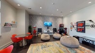 This single-floor amenity space is thoughtfully designed to blend entertainment, social gathering, and professional functionality in one cohesive environment. The main game room draws immediate attention with its bold textured accent wall in a neutral grey pattern, serving as a dynamic backdrop for a wall-mounted flat-screen TV and floating media console. Oversized bean bag chairs are arranged atop a vibrant multicolored area rug, creating a relaxed, casual seating zone perfect for gaming or lounging. A dedicated arcade racing simulator and a classic Pong table add to the entertainment offerings, while retro-inspired wall décor, including vintage-style signage and novelty plaques, lends the space a fun, eclectic personality. Dark hardwood flooring runs throughout, grounding the energetic design with a sleek, polished finish. A connected dining and activity area features round tables paired with bold red molded chairs, with wall-mounted shelves displaying board games — ideal for group gatherings. Natural light filters in through white-trimmed windows, brightening this social zone. Recessed lighting throughout ensures consistent, well-distributed illumination. Adjacent to the game room, a formal leasing or office area is accessed through tall glass-paneled double doors, revealing a striking teal botanical accent wall, a modern chandelier, and a clean, professionally appointed workspace that completes this well-rounded community facility.