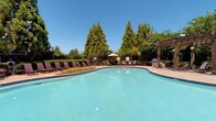 Located in Riverdale, Georgia, this single-story community amenity facility offers a well-rounded set of shared spaces designed for comfort and recreation. The centerpiece of the property is a generously sized outdoor swimming pool with a freeform shape and crystal-clear water, framed by a decorative brick paver surround. A substantial wooden pergola with overhead lighting anchors one side of the pool deck, providing shaded seating and an outdoor grill area ideal for gathering. Multiple lounge chairs line the poolside, and mature evergreen trees form a natural backdrop, enhancing privacy and the overall outdoor ambiance.

The adjacent single-story building features a neutral exterior with large windows that allow natural light to flow into the interior spaces. Inside, the common areas are finished with light-toned ceramic tile flooring throughout, contributing to a clean and practical aesthetic. A French-door entry leads to a fitness room equipped with cardio machines, visible through the glass panes. The interior corridors are finished with soft blue-gray walls, crown molding accents, and understated decorative touches such as framed artwork. Additional utility areas with tiled flooring and clean, functional layouts round out the interior. The overall atmosphere is practical, well-maintained, and community-oriented.