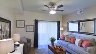 This single-story space showcases a well-appointed living room designed for both comfort and functionality. The room is anchored by a five-blade ceiling fan with an integrated light fixture, ensuring year-round comfort and ambient lighting. A large sliding glass door fitted with vertical blinds and dark accent curtains serves as the focal point of the space, allowing abundant natural light to fill the interior while providing privacy control.

The living area accommodates generous seating, including a plush sofa accented with decorative pillows in warm coral and floral tones, complemented by a coordinating armchair. A wooden coffee table with decorative accents centers the seating arrangement, grounded by a striped area rug that adds texture and definition to the space. A floor lamp provides additional warm lighting options.

Wall art and framed prints add character to the neutral-toned walls, while a framed mirror on the adjacent wall enhances the sense of openness. The combination of natural light from the sliding door, thoughtful furnishings, and layered lighting creates an inviting and practical living environment with a relaxed, contemporary aesthetic.