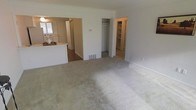 This single-floor residence offers a clean, functional layout designed to maximize livability and storage. The main living area is finished with wall-to-wall neutral carpeting, creating a soft and comfortable atmosphere throughout. An open passage connects the living space directly to the kitchen, which features hard flooring, white cabinetry, and a countertop peninsula that serves as a natural divider between the two areas. Natural light filters in through the kitchen window, brightening the shared space.

Toward the back of the living area, two closets are visible side by side — one featuring a standard hanging rod and the other outfitted with built-in shelving, offering versatile storage options for clothing, linens, or household essentials. A floor-level vent indicates a central heating and cooling system integrated into the unit.

The overall color palette is light and neutral, with white walls and trim throughout, contributing to a bright and airy feel. The straightforward, open design allows for flexible furniture arrangements, making the space adaptable to a variety of lifestyle needs. The seamless flow between the kitchen and living areas supports both everyday function and ease of movement across the single-floor layout.