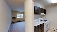 This single-floor residence offers a practical and comfortable layout designed for everyday living. The kitchen is equipped with dark wood cabinetry featuring arched door details, a gas range, and light granite-style countertops, providing ample storage and prep space. A structural column subtly separates the kitchen from the adjacent living area, creating a sense of definition between the two spaces without fully closing them off. The living area is carpeted and receives natural light through a multi-pane window, contributing to a bright and welcoming atmosphere. A baseboard heating unit runs along the wall beneath the window, and a window-mounted air conditioning unit ensures year-round climate comfort. The neutral wall tones throughout the space lend a clean, move-in-ready feel. The overall layout is efficient and well-suited for a single occupant or couple seeking a straightforward, low-maintenance living space with essential amenities conveniently within reach.