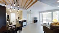 This single-floor space showcases a thoughtfully designed open-concept layout that integrates the living, dining, and kitchen areas into one cohesive environment. The interior is defined by striking exposed wood ceiling beams set against crisp white walls and ceilings, creating a warm industrial aesthetic. Polished concrete-style flooring runs throughout, lending a sleek, contemporary feel while enhancing the sense of continuity across the space.

The kitchen features a breakfast bar with bar seating, open shelving, and pendant Edison bulb lighting suspended from an artistic branching fixture, adding a stylish focal point overhead. A separate dining area accommodates a dark wood table and chairs, positioned conveniently adjacent to the kitchen. The living area is anchored by a media console and television, with a large sliding glass door or floor-to-ceiling window allowing abundant natural light to flood the room and providing access or views to the outdoors. A hallway visible from the main area suggests additional private rooms beyond. The overall ambiance is bright, airy, and modern, balancing industrial design elements with clean, minimalist finishes.