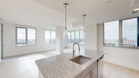 This thoughtfully designed 2-bedroom, 3-bathroom residence occupies a high floor, offering expansive panoramic city views through large floor-to-ceiling windows that flood every room with natural light. The open-concept kitchen and living area serves as the heart of the home, anchored by a central island finished with granite countertops, an undermount sink, and elegant pendant lighting. The kitchen is equipped with stainless steel appliances, including an electric range and built-in microwave, complemented by sleek two-tone cabinetry in white and gray. Light wood-tone flooring runs throughout, enhancing the bright, airy ambiance. The spacious living room features a corner window configuration, maximizing city views from multiple angles. A clean, well-lit hallway connects the private quarters, which include two generously sized bedrooms, each with large windows framing elevated urban vistas. The primary bedroom is well-proportioned and serene. All three bathrooms are finished with granite-topped vanities with blue-gray cabinetry, subway tile surrounds, and brushed gold fixtures. Two dedicated closets provide ample storage throughout the residence.