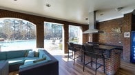 Located on Donerail Way in Raleigh, North Carolina, this single-level covered outdoor pavilion is designed to serve as a premier poolside gathering and entertainment destination. The structure showcases classic red brick construction, with large open archways that frame sweeping views of the adjacent swimming pool and surrounding mature tree canopy, allowing natural light and fresh air to flow freely throughout the space.

The pavilion is thoughtfully divided into distinct functional zones. A well-appointed outdoor kitchen and bar area anchors one side of the space, complete with dark granite countertops, a stainless steel range hood, and elevated bar seating with woven high-back chairs — ideal for casual dining and entertaining. The rich, warm-toned hardwood flooring runs throughout, lending a refined, cohesive aesthetic to the entire area.

Opposite the kitchen, a comfortable lounge area features plush wicker sectional seating with teal cushions and decorative throw pillows, providing an inviting spot to relax before or after a swim. Recessed ceiling lighting ensures the space remains functional into the evening hours. The open-air brick archways seamlessly connect the interior of the pavilion to the outdoor pool deck, creating a fluid indoor-outdoor experience perfectly suited to Raleigh's temperate climate.