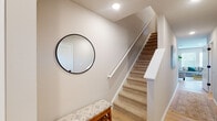 This two-floor home offers a thoughtfully designed layout with a clean, contemporary aesthetic throughout. The main level features light hardwood flooring that flows seamlessly through the living, dining, and kitchen areas. The living room is bright and welcoming, with multiple windows allowing generous natural light and views of the surrounding greenery. The kitchen is outfitted with white shaker cabinetry, dark granite countertops, a tile backsplash, stainless steel appliances, and a peninsula with bar seating — ideal for casual dining or entertaining. Adjacent to the kitchen, the dining area is anchored by a stylish glass-top table beneath a cluster of geometric pendant lights, with a large statement artwork adorning the wall. A dedicated laundry room with full-size washer and dryer adds everyday convenience on the main level. The staircase, carpeted in neutral tones with a clean white railing, leads to the upper level. The primary bedroom is generously proportioned, featuring plush carpet, vaulted ceilings, multiple windows, and a serene, light-filled atmosphere. The en-suite bathroom presents a sleek, modern design with large-format grey tile flooring, white cabinetry with gold hardware, a dark countertop, and a glass-enclosed walk-in shower — offering a refined, spa-like retreat.