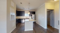 This single-floor residence offers a thoughtfully designed layout that balances functionality with modern style. The kitchen serves as the heart of the home, showcasing dark navy cabinetry that contrasts beautifully against light granite countertops. A central island with matching granite provides additional prep and dining space, illuminated by elegant pendant lighting. Stainless steel appliances — including a refrigerator, range, dishwasher, and microwave — complete this well-equipped kitchen, which is further enhanced by a classic subway tile backsplash and rich wood-look flooring.

The open living and dining areas flow naturally from the kitchen, featuring the same durable wood-look flooring, crown molding, and ceiling fans that lend a polished, cohesive feel throughout. Large windows with blinds invite natural light into every room, creating a bright and welcoming atmosphere.

The bedrooms offer plush carpeting underfoot, ceiling fans, and generous natural light from well-positioned windows. One bedroom provides direct access to an outdoor area through a glass-panel door, adding a convenient indoor-outdoor connection.

The bathroom is clean and functional, featuring a soaking tub with a tiled surround, a curved shower rod, wood-look tile flooring, and brushed nickel fixtures that tie the space together with a contemporary touch.