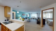 This single-floor residence offers a well-designed open-concept layout that connects the kitchen, living, and dining areas into one cohesive space. The kitchen is equipped with honey-toned oak cabinetry, white appliances, and a breakfast bar with seating, all set atop light hardwood flooring. A pendant light above the bar adds a decorative touch. The living area transitions seamlessly into the dining space, both finished with plush carpeting. A bold teal accent wall anchors the living room, which receives natural light through large windows dressed with sheer curtains. The dining area accommodates a bar-height table set near a sliding glass door that opens to a private balcony with outdoor views.

The primary bedroom features carpeted flooring, a textured ceiling, and direct balcony access through a sliding glass door. A second bedroom offers comparable comfort with carpet, a closet, and convenient proximity to an adjacent bathroom. Both bathrooms are appointed with wood-tone vanities, tile flooring, and tub-shower combinations. The overall ambiance is bright and functional, with warm wood trim accents throughout tying the design together cohesively.