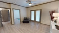 This single-story space is designed with a clean, contemporary aesthetic that emphasizes open, airy living. The main living area features light hardwood-style flooring that runs throughout, contributing to a cohesive and welcoming atmosphere. Neutral beige and greige walls are complemented by taupe-toned trim and crown molding, adding a subtle architectural detail that frames the room with a polished finish.

A ceiling fan with an integrated light fixture is centrally mounted, providing both ventilation and ambient lighting. French doors fitted with white horizontal blinds serve as a prominent feature, allowing natural light to filter into the space while offering privacy control and potential outdoor access. The doors create a bright focal point along one wall.

An open doorway connects to an adjacent room, also finished with matching flooring, suggesting a consistent design language carried throughout the space. The overall layout feels open and easy to navigate. With its understated color palette, practical fixtures, and light-enhancing features, the space presents a versatile and move-in ready environment suited to a variety of uses and furnishing styles.