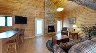 This log cabin showcases authentic rustic construction, with knotty pine tongue-and-groove walls and ceilings extending throughout every room, complemented by light hardwood flooring. Exposed log beams add structural character and reinforce the cabin's traditional craftsmanship.

The open living area serves as the heart of the cabin, anchored by a floor-to-ceiling stacked stone corner fireplace beneath soaring vaulted ceilings. Natural light filters in through multiple windows and glass-paneled doors, brightening the space while maintaining its cozy ambiance. The living room is furnished with plaid upholstered seating and a braided area rug, enhancing the rustic aesthetic.

The kitchen is well-appointed with light maple cabinetry, dark granite countertops, and a full suite of stainless steel appliances, including a refrigerator, dishwasher, range, and microwave. A granite-topped breakfast bar with wooden stools provides casual dining space.

An upper loft area, accessible via a staircase and overlooking the main living space, adds vertical dimension to the interior. A compact workspace features a built-in granite desk, computer setup, and a cork bulletin board, tucked neatly beneath rustic log ceiling beams.