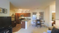 This single-story space showcases an open-concept layout that seamlessly connects the kitchen and dining area, creating a cohesive and functional living environment. The kitchen is well-appointed with rich, warm-toned cabinetry that extends to the upper walls, stainless steel appliances including a refrigerator, stove, and microwave, and a central island featuring a dark granite countertop with a built-in sink. Track lighting overhead illuminates the kitchen workspace effectively. The flooring transitions from hardwood-style planks in the kitchen to plush carpeting in the dining area, defining each zone while maintaining visual continuity. The dining area comfortably accommodates a round wooden table paired with industrial-style metal chairs, anchored beneath a pendant light fixture. Decorative wall art featuring Roman landmark typography adds a distinctive character to the space, complemented by a marquee-style illuminated "EAT" sign mounted above the kitchen cabinetry. A window allows natural light to filter into the space, enhancing the warm, inviting ambiance. The overall design blends modern functionality with eclectic, urban-inspired décor elements.
