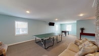 The Willowbrook community space is a thoughtfully designed single-floor layout offering a generous, open-concept environment ideal for gatherings and recreation. The interior is finished with soft blue-gray walls, neutral carpeting throughout most of the space, and recessed lighting that provides consistent illumination across the various activity zones.

A prominent brick column serves as the architectural centerpiece of the main area, visually dividing the lounge zone from the recreation zone while adding a warm, rustic contrast to the otherwise contemporary palette. The lounge area features plush leather sectional seating oriented toward a wall-mounted flat-screen television, creating a comfortable setting for socializing or viewing. French doors along one wall introduce natural light and provide access to an exterior exit.

The recreation area is generously sized to accommodate a full ping-pong table alongside a foosball table, with additional seating nearby. Multiple windows throughout the space bring in natural light from several directions.

A dedicated kitchenette offers dark espresso cabinetry, light granite-style countertops, a double stainless steel sink, and black appliances including a full-size refrigerator, providing functional support for events and gatherings. The overall ambiance is clean, open, and well-suited for a variety of community activities.