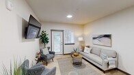 Located at 1500 Russell Ave SE in Port Orchard, Washington, this thoughtfully designed single-floor amenity space offers a well-rounded mix of social, culinary, and wellness facilities. The welcoming entry hallway features soaring vaulted ceilings, light hardwood flooring, and curated décor that sets a clean, modern tone throughout.

The open-concept living and dining area serves as the heart of the space. The living room is anchored by a striking white brick feature wall with a built-in electric fireplace and wall-mounted television, complemented by a circular pendant light and comfortable seating. Adjacent, the dining area offers a sleek communal table with modern leather chairs, connected to the living space through a full-length steel-framed mirror that enhances the sense of openness.

The kitchen is appointed with light wood cabinetry, white quartz countertops, a subway tile backsplash, stainless steel appliances, and open floating shelves — blending functionality with contemporary style. Terrazzo-style flooring adds a refined touch.

The dedicated exercise room is generously sized and outfitted with cardio machines, free weights, a cable machine, and a full-wall mirror. Exposed wood ceiling beams and large windows bring in natural light, creating an energizing environment for fitness. The space is well-suited for a variety of workout routines.