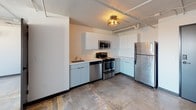 This single-floor unit offers an open, functional layout with an industrial character defined by exposed pipes, concrete-style flooring, and minimalist finishes throughout. The kitchen is a standout feature, designed in an L-shaped configuration with clean white cabinetry extending to the ceiling for ample storage. It is equipped with a full suite of stainless steel appliances, including a refrigerator, electric range, over-the-range microwave, and dishwasher, all set against light countertops. A ceiling-mounted drum light fixture adds a warm, modern accent to the space. The open living area is bright and simply finished, featuring white walls, a ceiling fan, and a wall-mounted mini-split HVAC unit for climate control. Exposed conduit runs along the ceiling, contributing to the unit's urban, loft-inspired character. The full bathroom includes a tub-shower combo with white tile surround, a toilet, and a vanity with a countertop, illuminated by an industrial-style flush-mount ceiling fixture. The consistent use of neutral tones and durable materials creates a cohesive, low-maintenance environment throughout the unit.