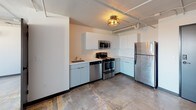This single-floor unit offers an open, functional layout with an industrial character defined by exposed pipes, concrete-style flooring, and minimalist finishes throughout. The kitchen is a standout feature, designed in an L-shaped configuration with clean white cabinetry extending to the ceiling for ample storage. It is equipped with a full suite of stainless steel appliances, including a refrigerator, electric range, over-the-range microwave, and dishwasher, all set against light countertops. A ceiling-mounted drum light fixture adds a warm, modern accent to the space. The open living area is bright and simply finished, featuring white walls, a ceiling fan, and a wall-mounted mini-split HVAC unit for climate control. Exposed conduit runs along the ceiling, contributing to the unit's urban, loft-inspired character. The full bathroom includes a tub-shower combo with white tile surround, a toilet, and a vanity with a countertop, illuminated by an industrial-style flush-mount ceiling fixture. The consistent use of neutral tones and durable materials creates a cohesive, low-maintenance environment throughout the unit.