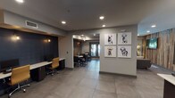 Located at 2121 Delgany St in Denver's vibrant LoDo neighborhood, this single-floor amenity space at The Metro Urban Apartments is a thoughtfully designed communal retreat. Guests are welcomed through glass-paneled entry doors bearing the property's logo, setting a polished first impression. Large-format gray tile flooring flows throughout, anchoring the open-plan layout that seamlessly connects multiple functional zones. The lounge area is a standout feature, centered around a reclaimed wood accent wall that adds warmth and texture, complemented by a wall-mounted flat-screen TV, tan leather armchairs, and a rustic wood coffee table. Floor-to-ceiling windows flood this seating area with natural light while offering views of outdoor greenery. Adjacent to the lounge, a dining and workspace area provides bistro-style tables and a built-in workstation with a desktop computer, ideal for residents needing a quiet spot to focus. The fully equipped kitchen features white subway tile backsplash, dark cabinetry, white quartz countertops, open shelving, stainless steel appliances, and a reclaimed wood accent wall echoing the lounge's aesthetic. Playful bulldog artwork adorns the walls, injecting personality into the space. Recessed lighting throughout ensures a warm, welcoming ambiance across all areas.
